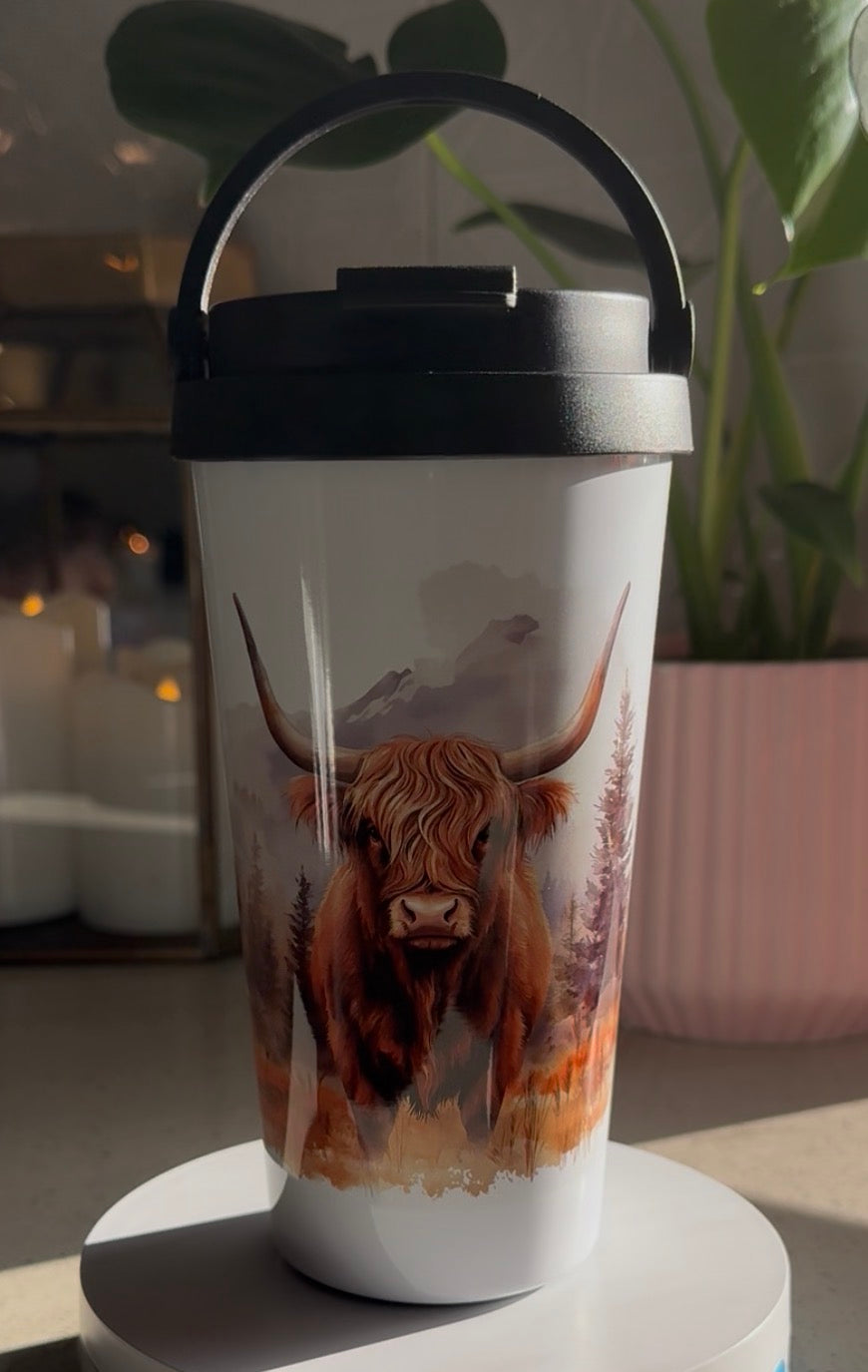 A white insulated travel tumbler with a black lid and carry handle, featuring a printed illustration of a Highland cow in a scenic landscape. The tumbler is displayed on a white round pedestal in a home setting with a blurred plant and candles in the background.