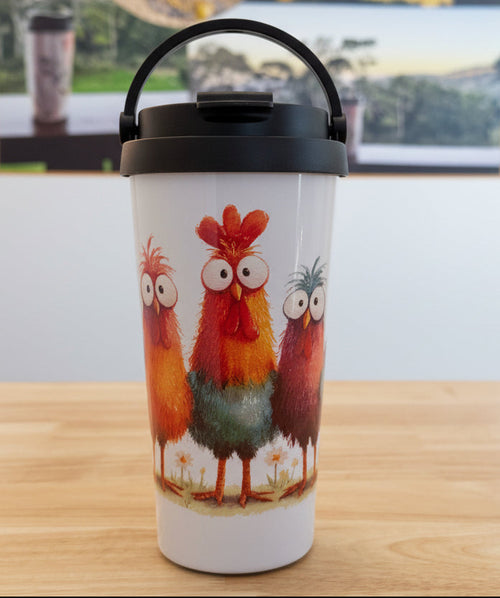 A white insulated travel tumbler with a black lid and handle, featuring a colorful cartoon rooster illustration, sitting on a wooden tabletop with a blurred product poster in the background.