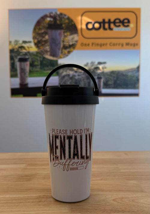 A white insulated travel tumbler with a black lid and carry handle sits on a wooden tabletop. The tumbler has the printed phrase 'PLEASE HOLD I'M MENTALLY Buffering' in black and brown lettering.