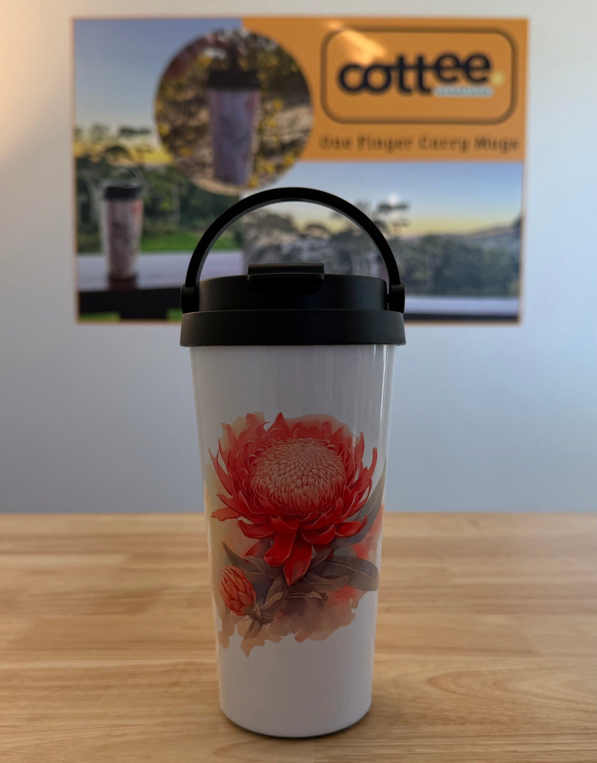 A white insulated travel tumbler with a black lid and carry handle, featuring a red and orange floral print, sitting on a wooden tabletop with a coffee-themed poster in the background.