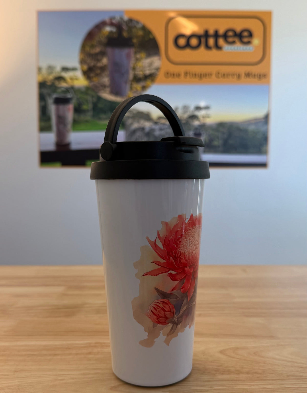 A white travel tumbler with a black lid and handle, featuring a red and orange floral graphic, sitting on a wooden table with a blurred 'cottee' sign in the background.