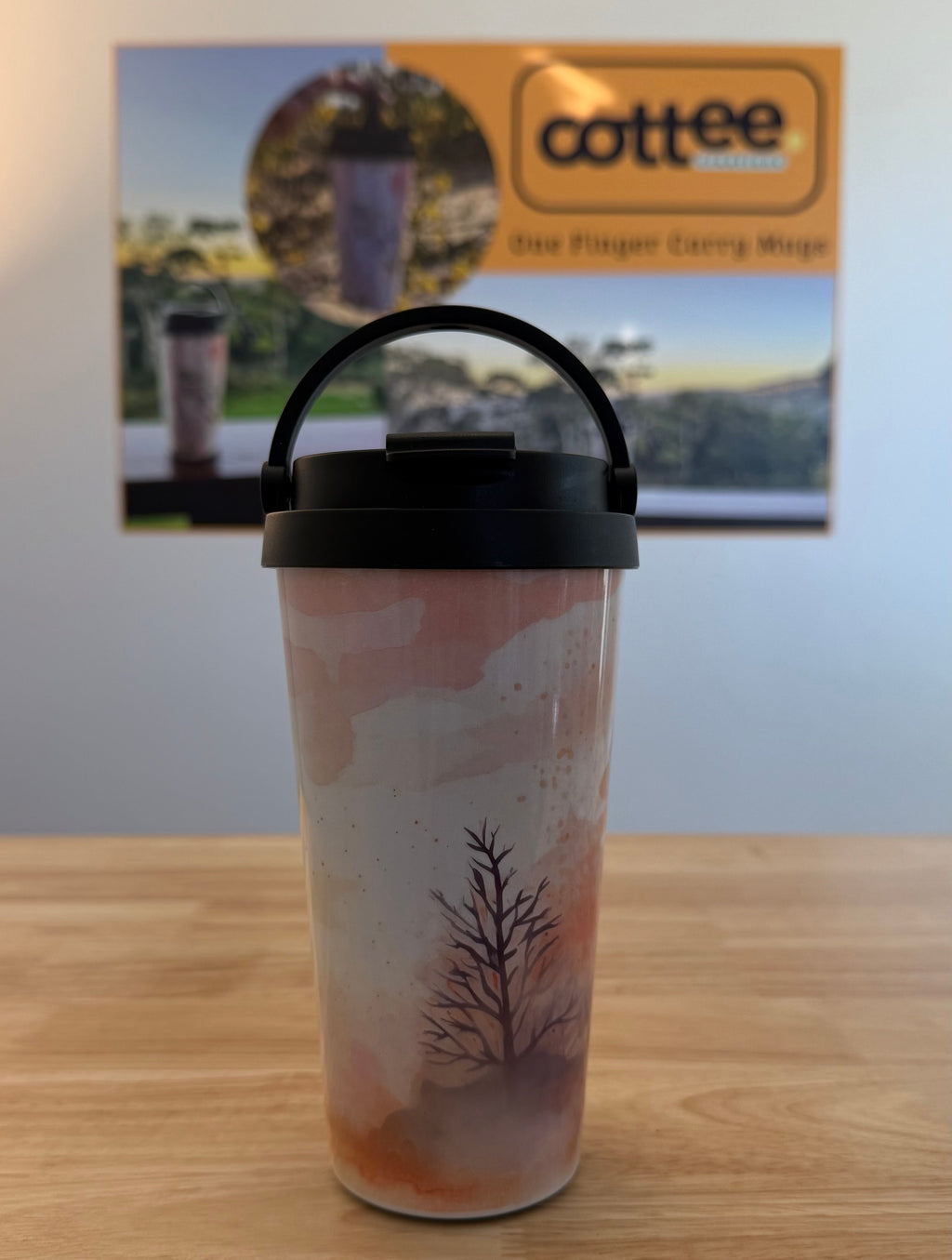 A travel tumbler with a black handle lid sits on a wooden table. The tumbler has a pink and white watercolor-style design with a dark tree illustration.