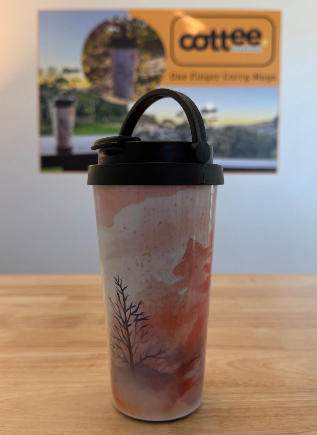 A travel tumbler with a black lid and carry handle sits on a wooden table. The tumbler has a printed design of a tree in a misty forest with orange, pink, and gray tones.