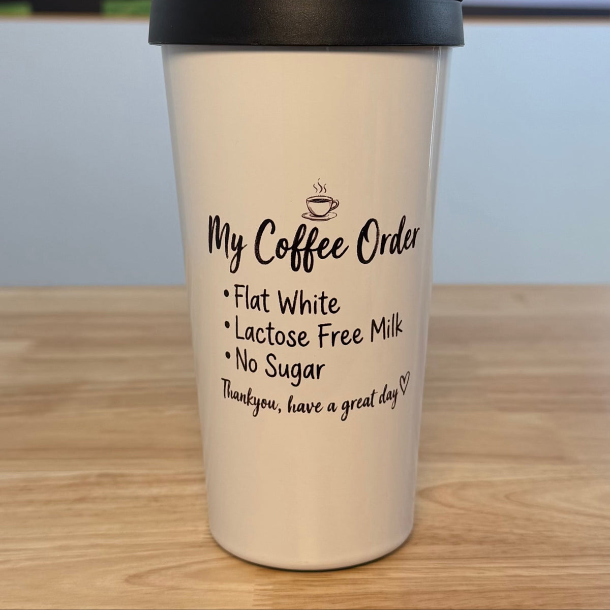 A white insulated travel tumbler with a black lid and carrying handle, sitting on a wooden tabletop. The tumbler features a black printed text design reading 'My Coffee Order' followed by a list of coffee preferences and a closing phrase.