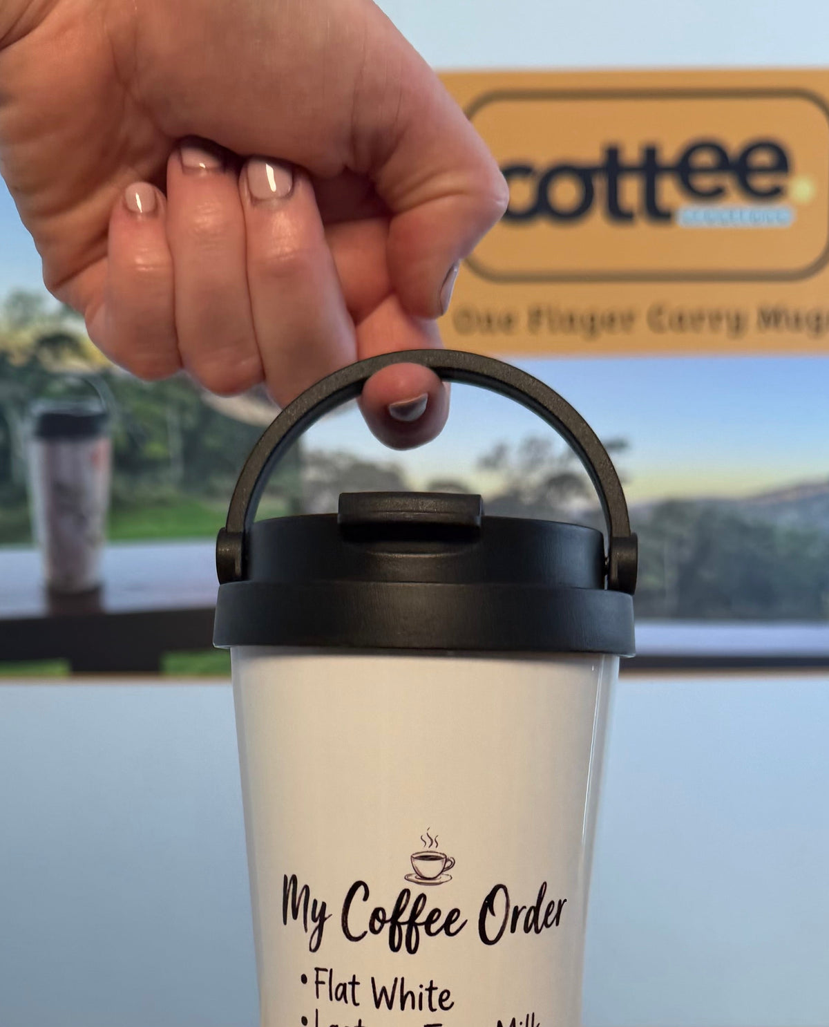 A hand holds a white insulated travel tumbler with a black lid and carry handle, printed with the text 'My Coffee Order' and a coffee cup graphic, on a light wood tabletop with a blue wall and a coffee sign in the background.