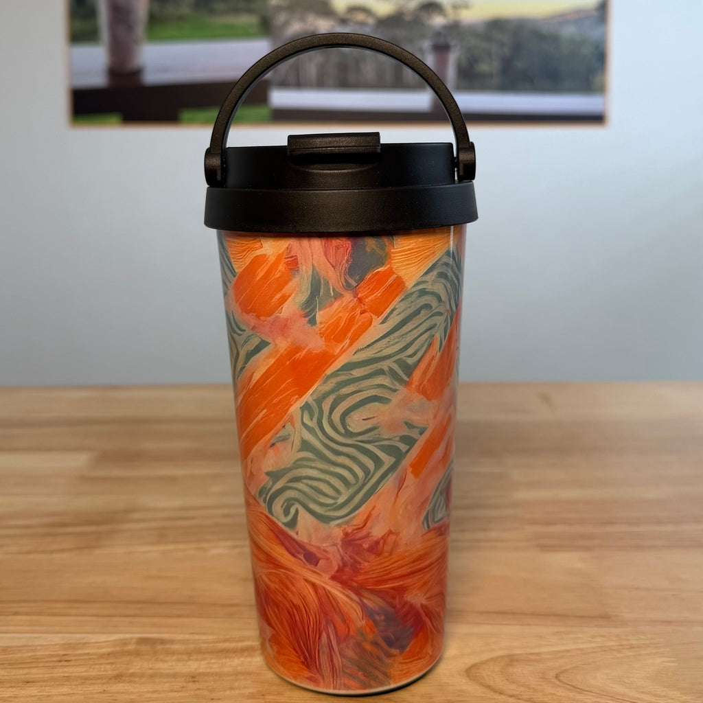 A travel tumbler with a black lid and handle, featuring an orange and green abstract pattern, sitting on a wooden table with a coffee poster in the background.