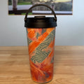 A travel tumbler with a black lid and handle, featuring an orange and green abstract pattern, sitting on a wooden table with a coffee poster in the background.