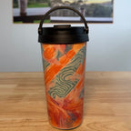 A travel tumbler with a black lid and handle, featuring an orange and green abstract pattern, sitting on a wooden table with a coffee poster in the background.