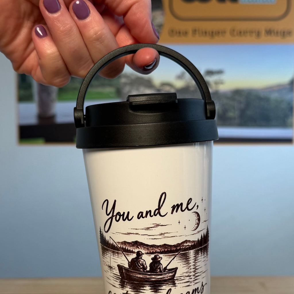 A hand with purple nail polish holds a white travel tumbler with a black lid and handle. The tumbler features a black illustration of a couple in a boat on a lake with the text 'You and me casting dreams at twilight'.