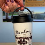 A hand with purple nail polish holds a white travel tumbler with a black lid and handle. The tumbler features a black illustration of a couple in a boat on a lake with the text 'You and me casting dreams at twilight'.