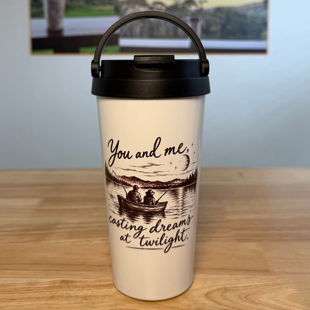 A white insulated travel mug with a black lid and handle sits on a wooden tabletop. The mug has a printed illustration of a person in a boat on water with the text 'You and me casting dreams of twilight.