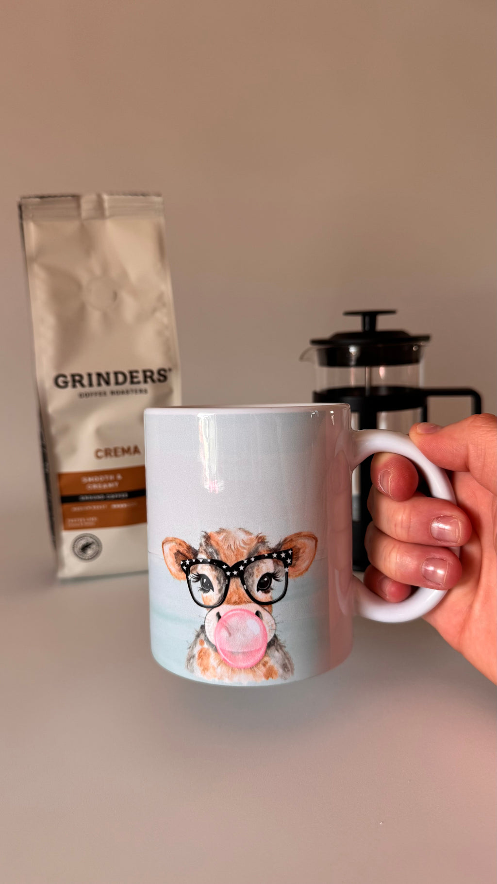 Mug with a cow design holding coffee, grinder, and coffee bag in the background.