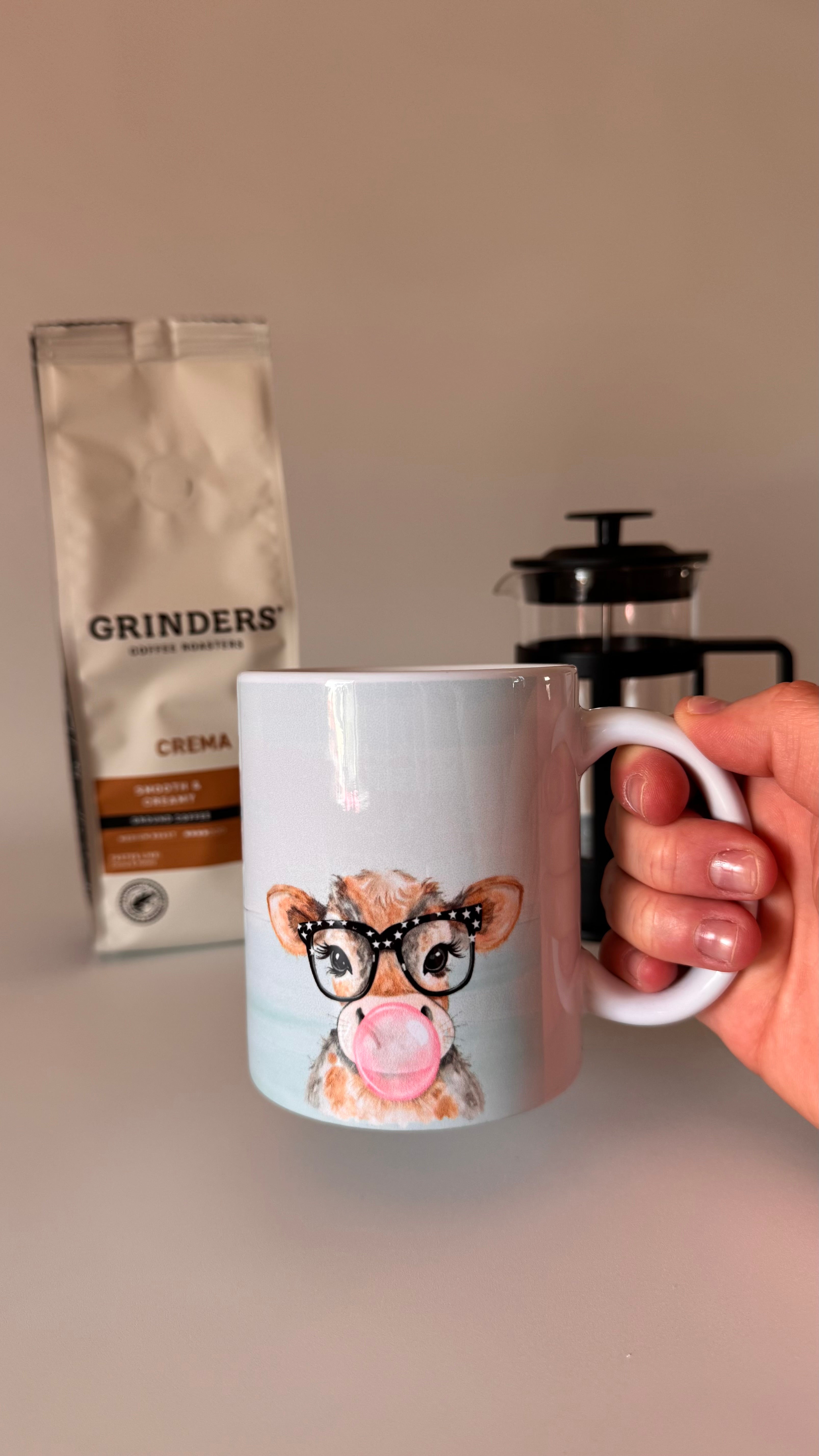 Mug with a cow design holding coffee, grinder, and coffee bag in the background.