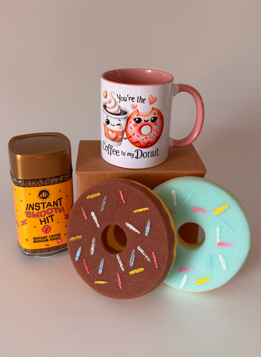 Mug with cartoon donuts, instant coffee jar, and two donut-shaped sponges on a neutral background