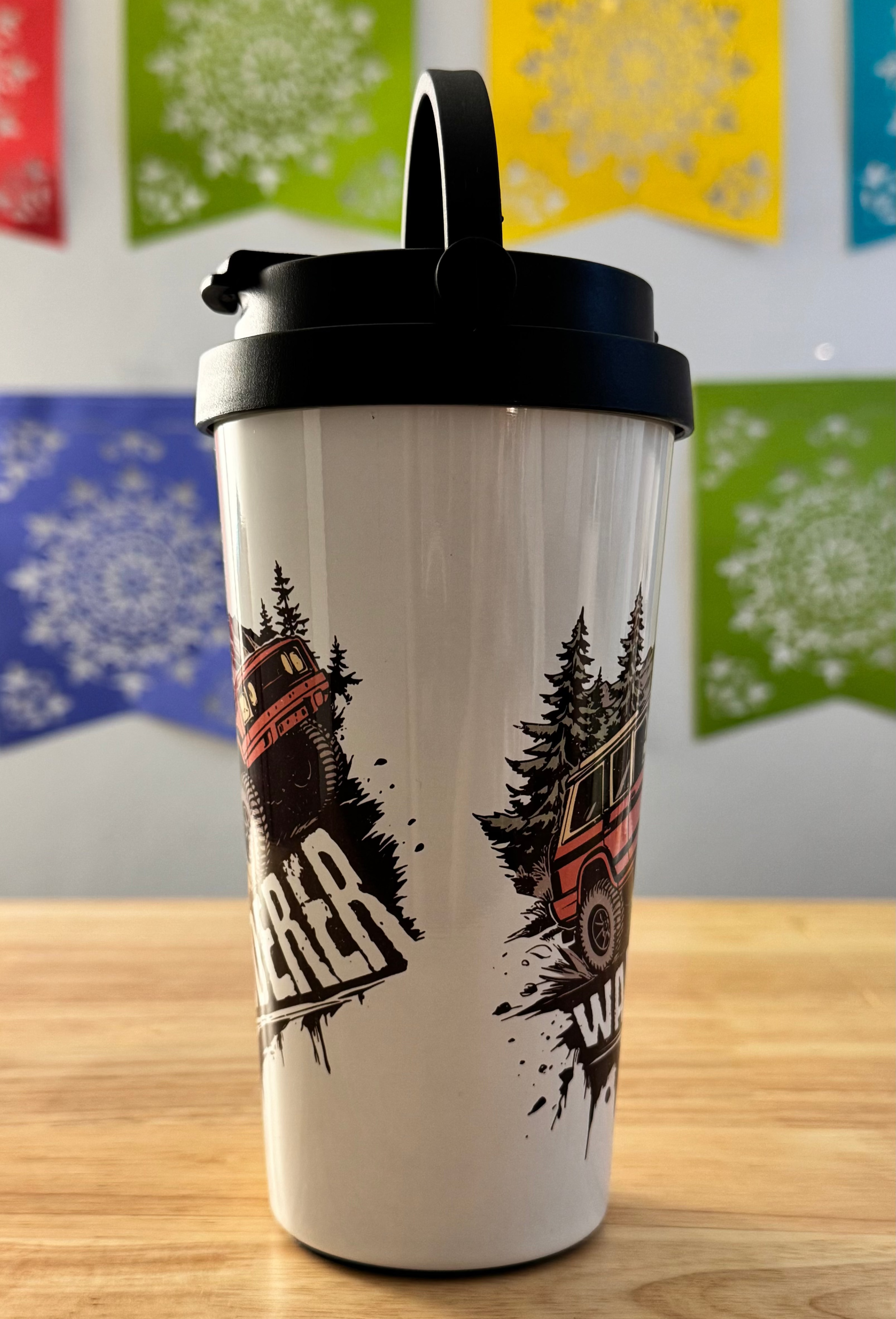 White tumbler with black lid featuring a graphic design of a truck and trees on a wooden surface with colorful flags in the background.