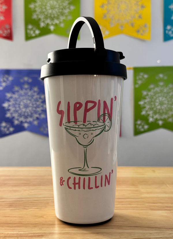 Tumbler with 'Sippin' & Chillin'' text and a cocktail graphic on a wooden surface with colorful flags in the background.