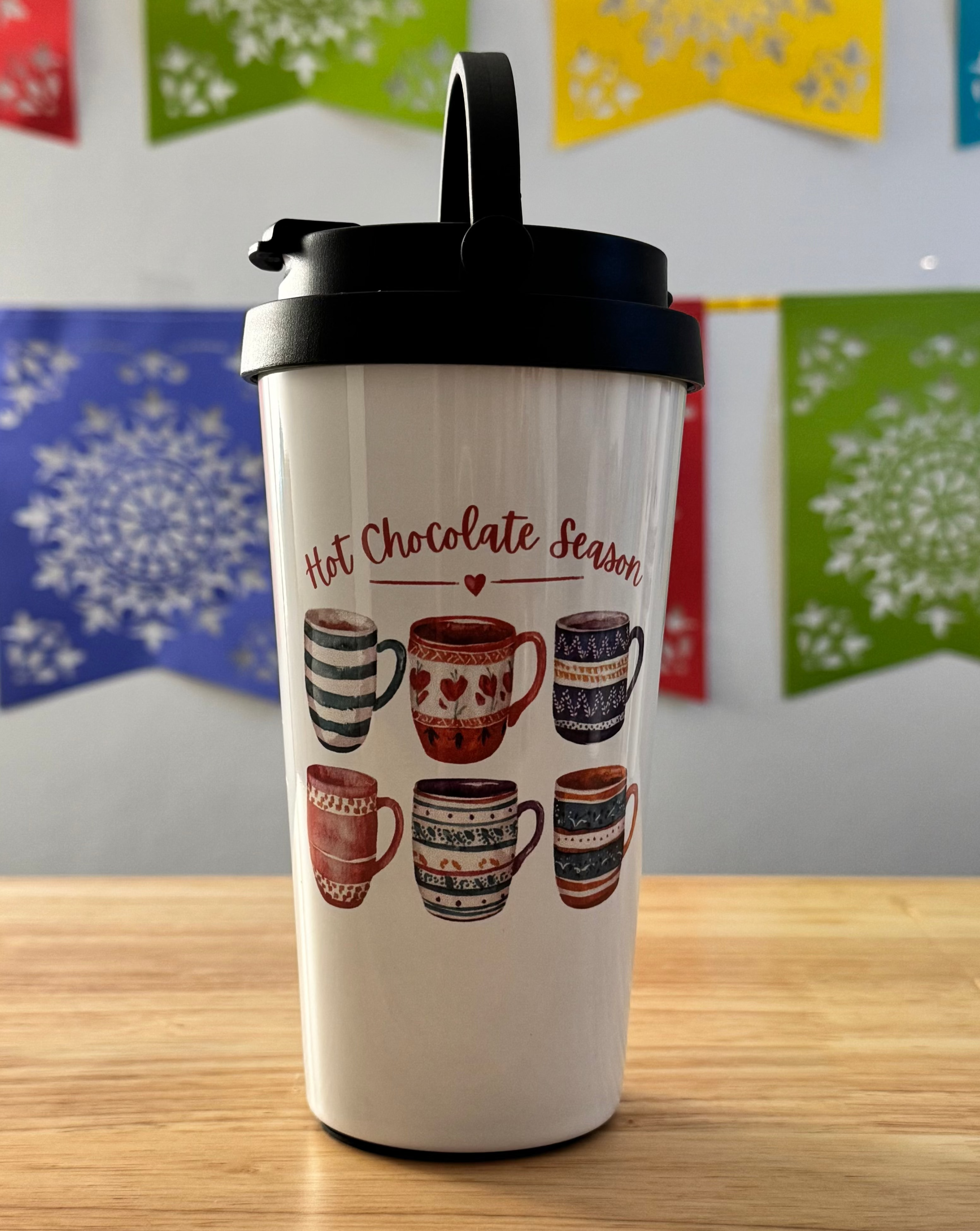 White tumbler with hot chocolate-themed design on a wooden surface with colorful snowflake decorations in the background.