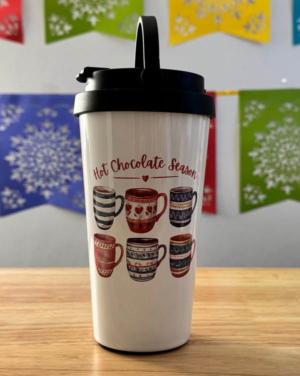 White tumbler with hot chocolate-themed design on a wooden surface with colorful snowflake decorations in the background.
