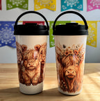 Two travel mugs with highland cow designs on a wooden surface with colorful flags in the background.