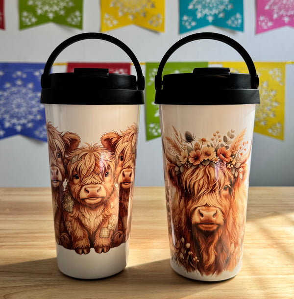 Two travel mugs with highland cow designs on a wooden surface with colorful flags in the background.