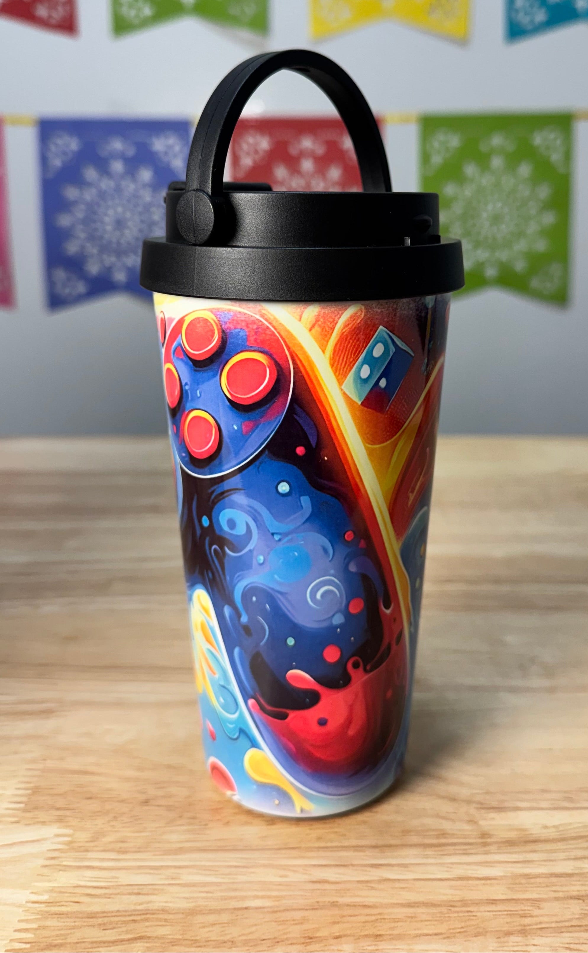 A travel tumbler with a black lid and carry handle, decorated with a colorful cosmic design of swirling blue, red, orange, and yellow patterns, set on a wooden table with a festive bunting banner in the background.