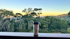 A reusable insulated tumbler with a floral print and black lid sits on a wooden railing, overlooking a scenic forest landscape at sunset.