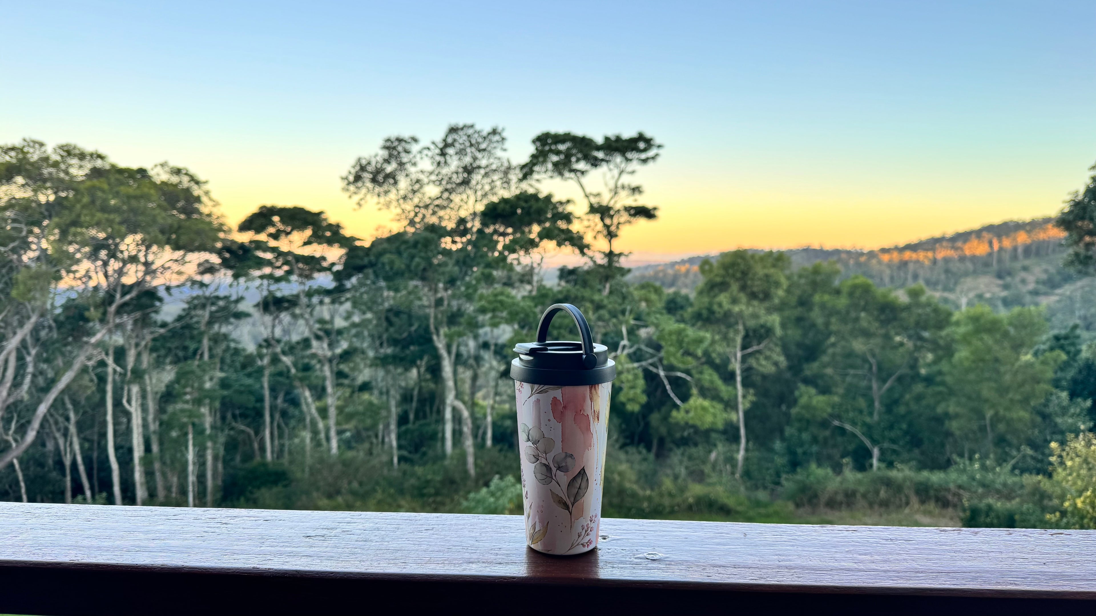 A reusable insulated tumbler with a floral print and black lid sits on a wooden railing, overlooking a scenic forest landscape at sunset.