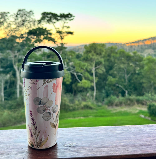 A travel tumbler with a floral pattern and black lid sits on a wooden railing with a scenic outdoor landscape and sunset in the background.